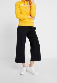 Sweat à capuche jaune avec logo blanc, cordon de serrage et poignets côtelés ; associé à un pantalon large noir avec une poche latérale et des boutons à l'ourlet ; baskets blanches.