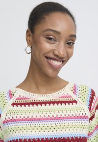 Usmievavá žena s tmavými vlasmi stiahnutými dozadu, nosí strieborné kruhové náušnice a farebný pruhovaný háčkovaný top na bielom pozadí.