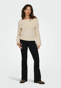 Sweter w kolorze kremowym, odsłaniający ramiona, z fakturowanym wzorem, prążkowanymi mankietami oraz dopasowanym krojem, w zestawie z czarnymi dzwonowymi dżinsami i brązowymi botkami za kostkę.