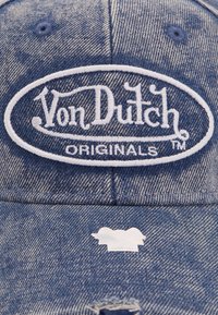 Denim kapa v zbledele modri barvi, z belim vezenim ovalnim logotipom z napisom "Von Dutch Originals." Vključuje majhno oznako na šiblji.