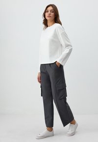 Weißes Langarmoberteil mit dekorativen Strasssteinen, kombiniert mit dunkelgrauen Cargo-Hosen mit Seitentaschen. Weiße Sneaker vervollständigen das Outfit.