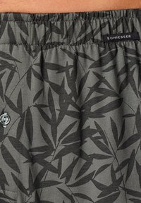 Boxers gris avec un motif de feuilles noires, taille élastique et fermeture à bouton à l'avant. Le tissu semble doux avec une finition légèrement texturée.
