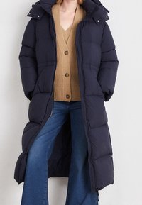 Marineblauer gesteppter Steppmantel mit Kapuze, mit gestepptem Muster und Druckknöpfen. Getragen über einer beige gestrickten Strickjacke und Jeans mit ausgestelltem Bein.