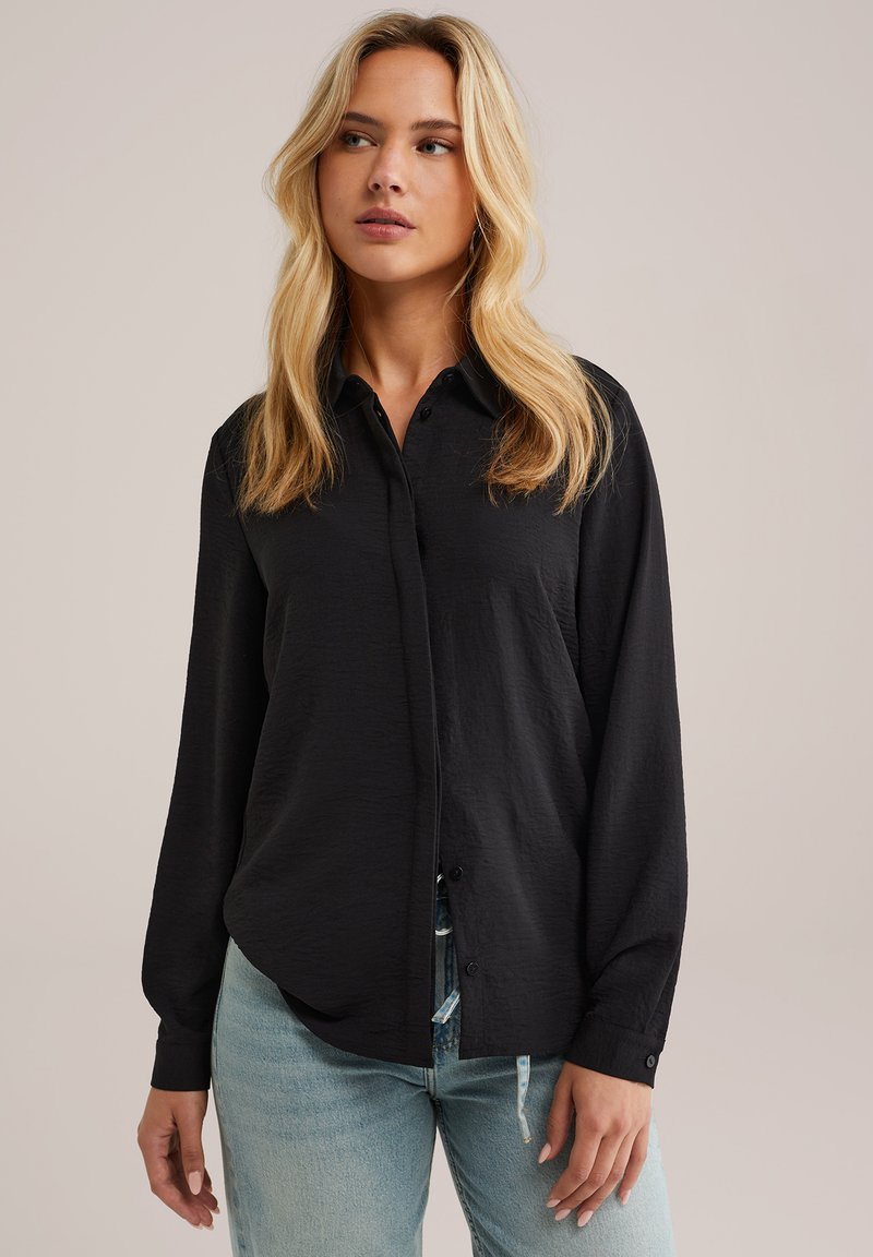 Donna bionda con capelli ondulati, che indossa una camicia nera a maniche lunghe abbottonata e jeans azzurri chiari, che guarda leggermente verso sinistra.
