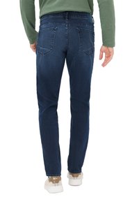 Jeans en denim bleu foncé avec une coupe ajustée, dotés de deux poches arrière et de coutures discrètes. Portés avec des baskets blanches et un haut vert.