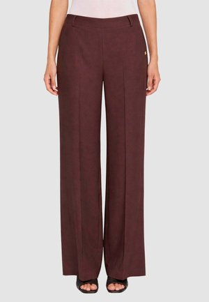 Donna che indossa pantaloni a vita alta e gamba larga di colore marrone scuro abbinati a tacchi neri aperti, mostrata dalla vita ai piedi su uno sfondo semplice.