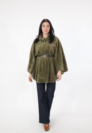 Kvinde iført en olivengrøn bæltejakke af kunstpels, mørkeblå jeans og brune sko, stående mod en hvid baggrund.