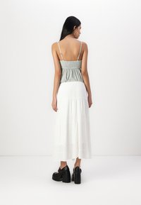 La robe maxi présente un haut gris froncé et une jupe blanche à volants. Le mannequin porte des chaussures plateforme noires et des boucles d'oreilles créoles. Vue arrière montrée.