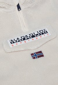 Krämfärgad fleece-pullover med halvmuddsdragkedja framtill, med en logotypapplikation och en liten norsk flagga på bröstet. Mjuk textur.