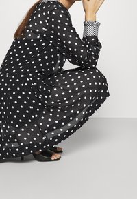 Robe noire transparente à pois blancs, manches longues, et col à carreaux. Associée à des sandales noires à bouts ouverts.