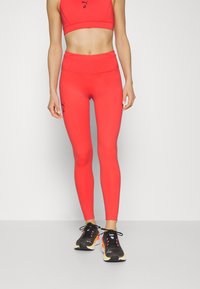 Czerwone dopasowane legginsy z wysokim stanem, wykonane z elastycznej tkaniny, zestawione z pasującym biustonoszem sportowym i czarnymi butami sportowymi.