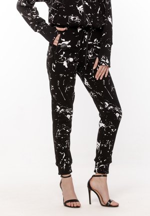 Czarne spodnie z białym wzorem rozprysków farby, z kieszenią na suwak z przodu, ściągaczem w pasie, noszone z czarnymi sandałami na wysokim obcasie i pasującą kurtką.