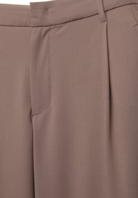 Gros plan sur un pantalon taupe montrant la taille, les passants de ceinture, le pli avant et le détail de la poche latérale.