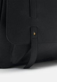 Sac en cuir noir avec un rabat à bords cousus et une sangle maintenue par un rivet en laiton, reposant sur une surface blanche.