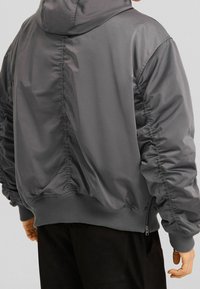 Grå huva jacka med en slät, glänsande yta, samlade sidodetaljer och ribbade muddar och fåll. Sidofickan med dragkedja är synlig.