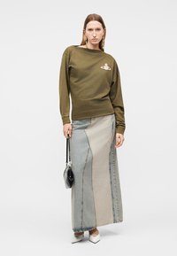 Γυναίκα με μακρυμάνικη μπλούζα ελιάς με έμβλημα, μάξι φούστα τζιν με πατζάκια, λευκά μυτερά παπούτσια, κρατώντας μια μικρή μαύρη τσάντα.