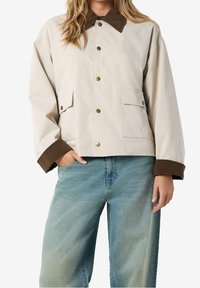 Kvinde iført en beige knappet jakke med brunt krave og manchetter, blå brede jeans, og den ene hånd i lommen.
