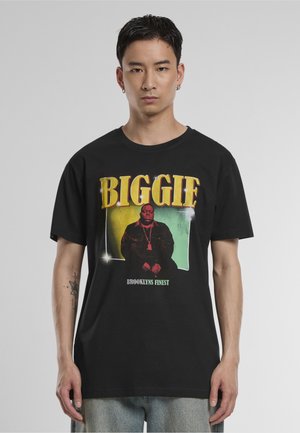 Junger Mann mit kurzen dunklen Haaren, der ein schwarzes T-Shirt mit einem farbenfrohen Porträt und dem Text "BIGGIE" und "BROOKLYN'S FINEST" trägt.