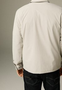 Lichtgrijs shirt met lange mouwen, voorzien van een rugstuk en manchetten met knopen. Heeft een zachte textuur en een ontspannen pasvorm, gecombineerd met donkere spijkerbroeken.