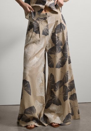 Pantalon beige à jambes larges avec de grands motifs de feuilles noir et blanc, porté avec un haut assorti et des sandales ouvertes.