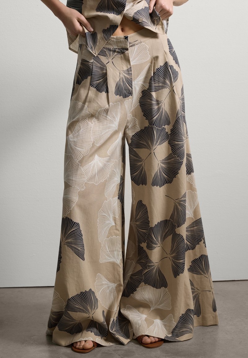 Pantalon beige à jambes larges avec de grands motifs de feuilles noir et blanc, porté avec un haut assorti et des sandales ouvertes.
