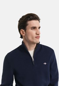 Mornarica pulover s kratkim zadrgo, izdelan iz teksturirane pletenine, z značilno srebrno zadrgo in malim logotipom na prsih. Klasičen dizajn, prilegajoč slog.