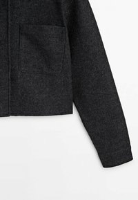 Dunkelgraue, kurz geschnittene Jacke aus strukturiertem Wollstoff, mit einer einzelnen Brusttasche und langen Ärmeln mit umgeschlagenen Enden.