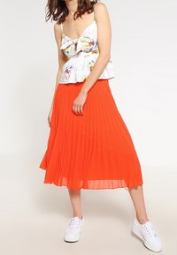 Bílé květinové top s peplum střihem a detailem mašle, spojený s plisovanou oranžovou sukní a bílými teniskami. Lehčí materiál a živé barvy.