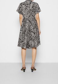 Frau, die ein knielanges, schwarz-weißes Kleid mit Blumenmuster und kurzen Ärmeln trägt, sowie schwarze, Absatzsandalen, steht vor einem schlichten Hintergrund.