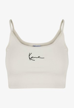 Světle béžový crop top s tenkými ramínky a přiléhavým střihem. Na přední straně se nachází vyšívaný černý podpis. Hladká textura a elastický materiál.