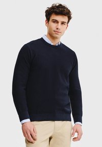 Granatowy sweter z dzianiny z okrągłym dekoltem i ściągaczami. Noszony na jasnoniebieskiej koszuli z kołnierzykiem, w zestawieniu z beżowymi spodniami.