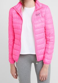 Rosa puffjacka med quiltat mönster, full dragkedja och hög krage. Har en liten logotyp på vänster bröst och matchande dragkedjeinslag.