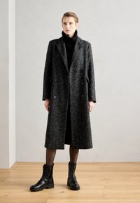 Schwarzer und grauer, gesprenkelter Wollmantel mit einem langen, geraden Schnitt, großen Revers, doppelter Knopfleiste und Seitentaschen. Kombiniert mit robusten schwarzen Stiefeln.