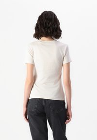 Beige short-sleeve t-shirt with a round neckline, smooth cotton texture. Paired with dark denim jeans, viewed from the back.