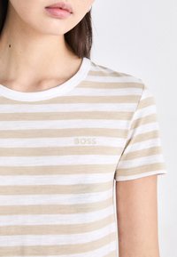 T-shirt beige et blanc rayé avec un col rond et des manches courtes. Présente un subtil logo "BOSS" de couleur assortie. Matière en coton doux.