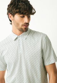 Lysegrå polo-shirt med et subtilt mønster over det hele, korte ærmer og standard krave. Stoffet ser glat og let ud.
