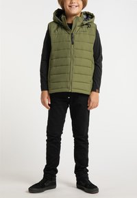 Gilet imbottito verde oliva con cappuccio, chiusura frontale con zip e coulisse elastiche. Indossato sopra una maglietta a maniche lunghe nera e pantaloni neri.