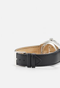 Reloj de pulsera de cuero negro con hebilla de metal plateado, que presenta una caja redonda y una correa de textura suave. El forro es de color marrón claro.