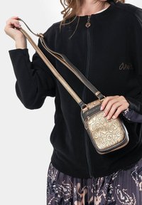 Metallic gold Umhängetasche mit geprägt gestalteter Schrift, schwarzen Akzenten, verstellbarem Riemen und Reißverschluss, gehalten von einer Person in einer schwarzen Jacke.
