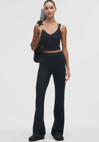 Marineblå crop top og flagrende bukser lavet af blødt stof. Modellen holder en yogamåtte og har en sort rygsæk med én strop.