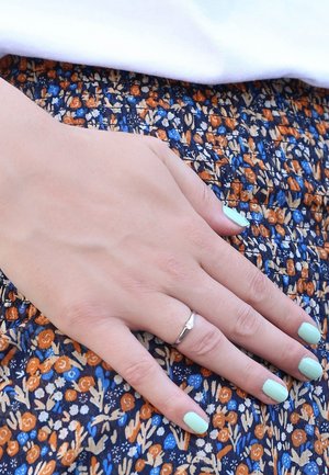 Zilveren ring op een hand met muntgroene nagellak, rustend op een stof met bloemenpatroon in oranje, blauw en crème kleuren.