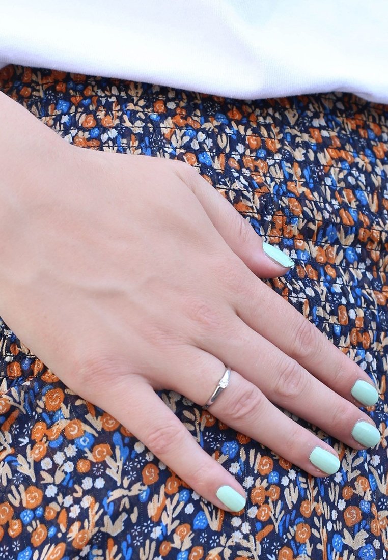 Zilveren ring op een hand met muntgroene nagellak, rustend op een stof met bloemenpatroon in oranje, blauw en crème kleuren.