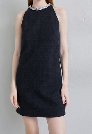 Czarna, bezrękawnikowa mini sukienka z fakturowanego materiału z ozdobnym srebrnym kołnierzem z koralików, noszona przez stojącą osobę na tle szarej ściany.