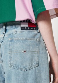 Světle modré džíny z denimu s nášivkou Tommy Jeans na pase a malým logem na zadní kapse, nošeny s zelenou a růžovou košilí.