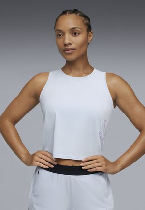 Frau mit geflochtenem Haar, die ein ärmelloses weißes Crop-Top und weiße Shorts mit schwarzem Taillenbund trägt, steht mit den Händen in der Hüfte.