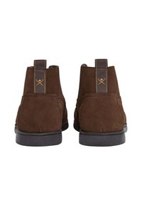 Para brązowych zamszowych botków za kostkę z czarnymi podeszwami, przedstawionych z tyłu, z haftowanymi paskami do zakupu i żółtym emblematem skrzyżowanych kijów golfowych.