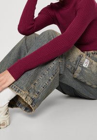 Pruhované burgundské triko s dlouhým rukávem v kombinaci s volnými, bledě modrými džíny s kapsou na boku a nedokončeným lemem. Viditelné bílé tenisky.