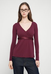 Νεαρή γυναίκα με μακριά καστανά μαλλιά, φορώντας μαύρα γυαλιά, μπορντό τοπ με σατέν ζώνη και σκούρο μπλε τζιν, στέκεται μπροστά σε απλό φόντο.