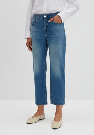 Vrouw die een lichtgekleurde, witgeknoopte blouse draagt, gecombineerd met een hoge taille, rechtgesneden blauwe spijkerbroek met vervaagde delen, en off-white platte schoenen, staande tegen een effen achtergrond.