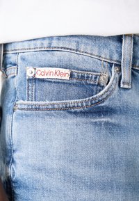 Világoskék farmernadrág Calvin Klein címkével, klasszikus szabással, elülső zsebeléssel és rézszínű varrási díszítéssel.